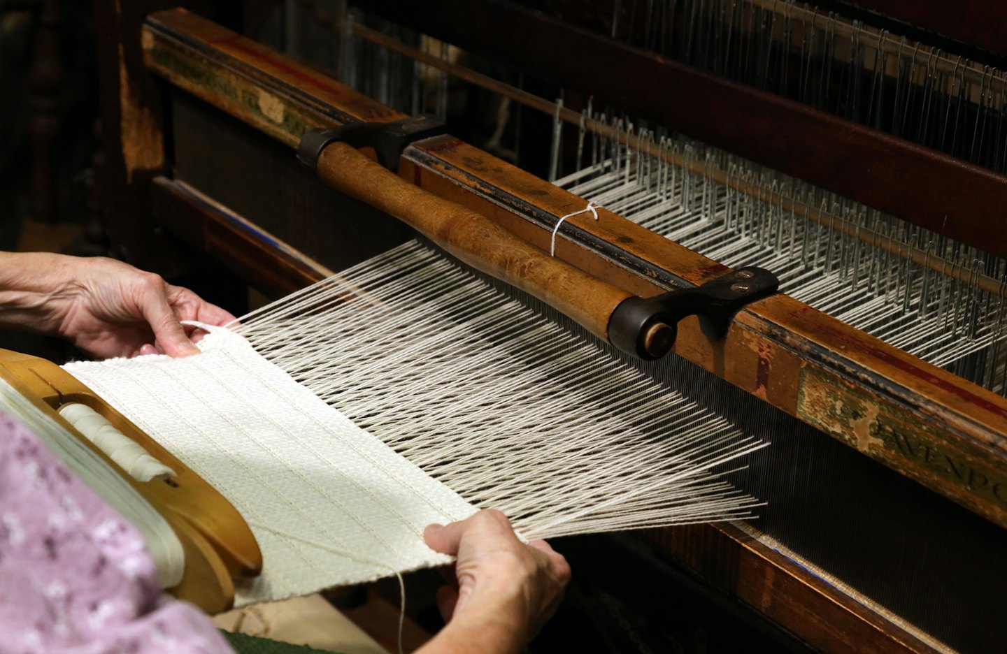 Wrinkled hands weaving white thread on a wooden loom.